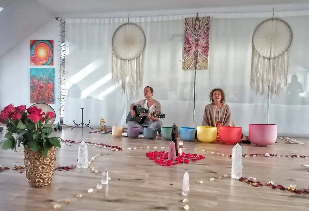 Kryštálové spievajúce misy rozložené v rade medzi sviečkami a lupeňmi ruží, vzadu sedia dvaja ľudia pri svetlých závesoch.